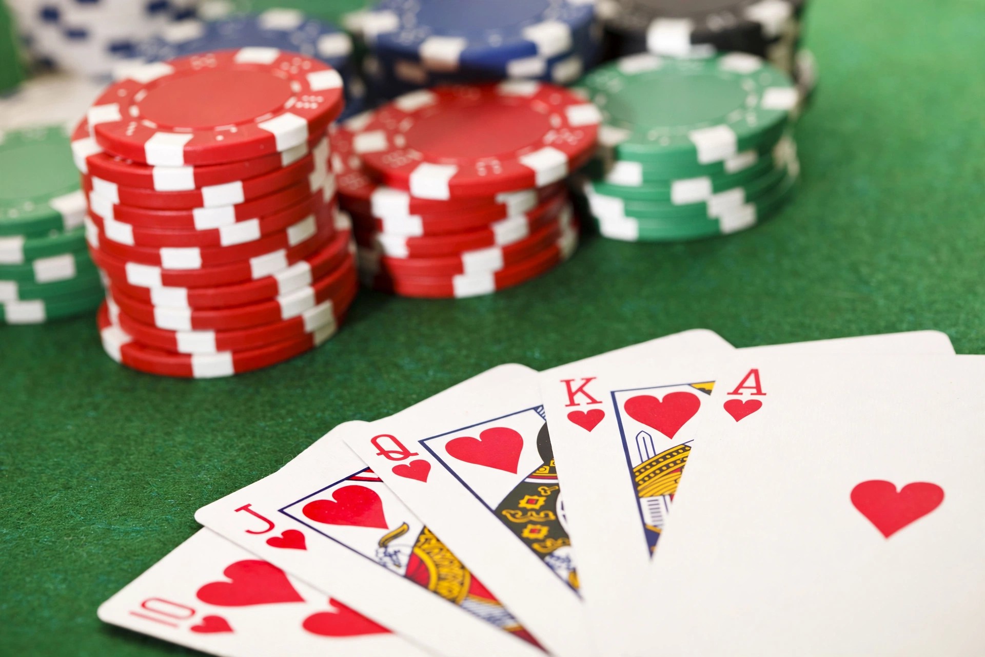 Stacks of red, green, blue and black poker chips on green felt beside playing cards fanned face-up showing a royal flush in hearts.